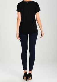 Camisa negra de manga corta con cuello redondo, combinada con jeans ajustados oscuros y zapatos de tacón alto negros, vista desde atrás.