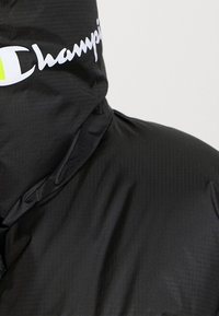 Schwarze Kapuzenjacke aus glänzendem Material mit horizontaler Textur. Verfügt über ein auffälliges weißes "Champion"-Logo auf der Kapuze.