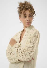 Blouse beige clair avec manches longues en dentelle à motif floral, col mao et coupe décontractée. Texture lisse avec un tombé doux.