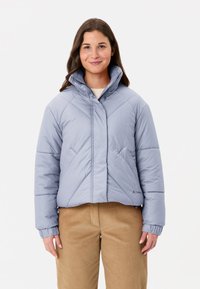 Giacca imbottita azzurro chiaro con colletto alto, cuciture diagonali e polsini elastici. Abbinata a pantaloni di velluto a coste beige.