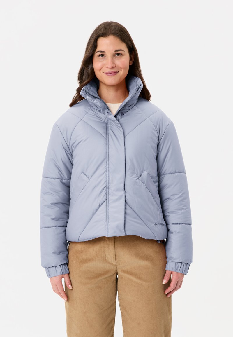 Giacca imbottita azzurro chiaro con colletto alto, cuciture diagonali e polsini elastici. Abbinata a pantaloni di velluto a coste beige.