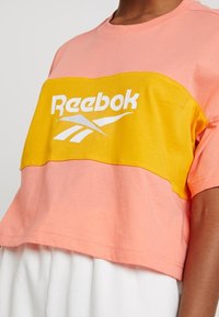 Zkrácený tričko s růžovou horní částí a žlutou dolní částí. Na přední straně je bílé logo Reebok. Měkký bavlněný materiál.