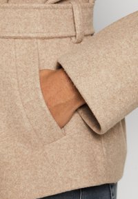 Μπεζ ζακέτα από μείγμα μαλλιού με υφαντή υφή, που διαθέτει τσέπη και μανσέτα που επεκτείνεται πέρα από τον καρπό, προσφέροντας κομψή εμφάνιση.