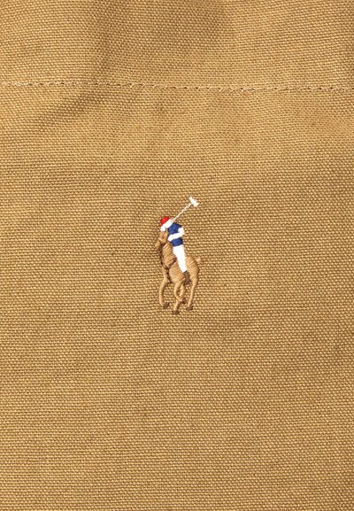 Κεντημένος παίκτης πόλο με κόκκινο κράνος, μπλε πουκάμισο, λευκό παντελόνι και υψωμένο μπαστούνι πόλο σε υφασμάτινο υπόβαθρο μπεζ με υφή.