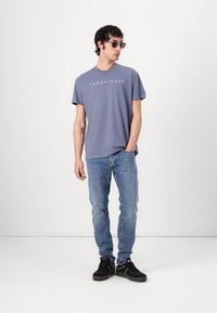 Rövid ujjú, világoskék pamut póló "TOMMY JEANS" logóval. Közepes mosású farmernadrággal és fekete sportcipővel párosítva. Kiegészítők között napszemüveg is szerepel.
