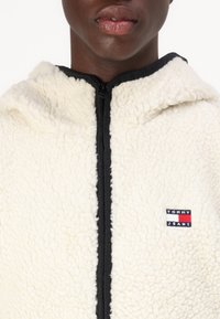 Vit fleece-hoodie med texturerad yta, svart dragkedja och broderad 'TOMMY JEANS'-logotyp på vänster bröst. Med huva.