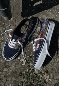 Een paar Vans sneakers met kleurrijke pluizige stof en metalen spikes, witte veters los, geplaatst op ruwe aarde met kleine planten in de buurt.