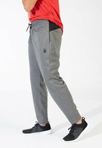 Grijze sportbroeken met een gestructureerde afwerking, zwarte zijpaneel, elastische tailleband en een klein logo op de linkerdij. Gecombineerd met zwarte schoenen.