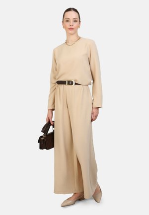 Combinaison beige à manches longues et ceinture. Tissu lisse, jambes larges et fente sur le côté. Associée à un sac à main marron et des ballerines claires.