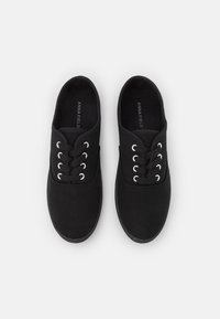 Czarne sneakersy z płótna z okrągłym czubkiem, sześcioma otworami na sznurówki z metalowymi oczkami oraz teksturowaną podeszwą. Nazwa marki widoczna wewnątrz.