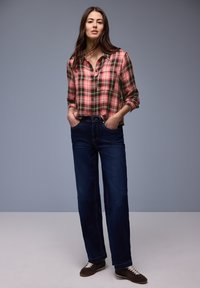 Femme debout, les mains dans les poches, portant une chemise à carreaux rose et verte, un jean bleu foncé et des chaussures marron foncé à lacets, sur fond gris.