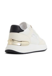 Weißer Sneaker mit schwarzen Akzenten, strukturierten Einsätzen und einem metallisch goldenen Detail an der Ferse. Dicke, gepolsterte Sohle für Unterstützung.