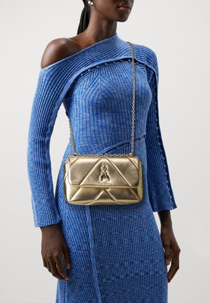 Sac à épaule matelassé doré avec une bandoulière en chaîne, arborant un emblème de marque à l'avant. Le matériau a une finition métallique brillante.