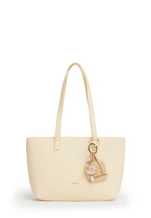 Crèmekleurige totebag met lange hengsels en een klein geweven bedeltje met schelp- en kralendecoraties dat aan één hengsel hangt.