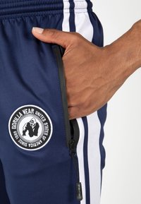 Marineblauwe sportbroek met witte zijkanten, voorzien van een ritszak en een rond logo met daarop een gorilla. Glad oppervlak, duurzaam materiaal.