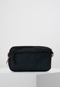 Rektangulär marinblå väska i tyg; har en framficka med dragkedja, svarta detaljer och en liten brun accent på ena sidan.