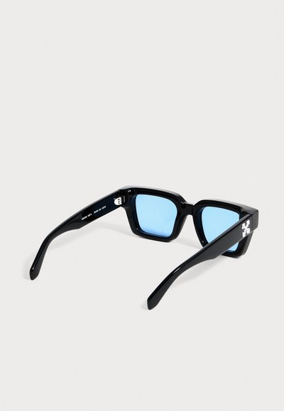 Lunettes de soleil rectangulaires noires avec des verres bleu clair et un logo blanc sur les branches, présentées sur un fond uni.