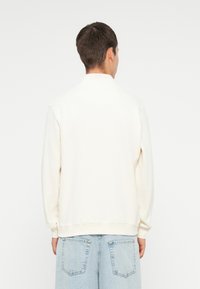 Felpa color crema con collo alto e polsini a costine. Vestibilità comoda con una texture liscia, abbinata a jeans baggy blu chiaro.