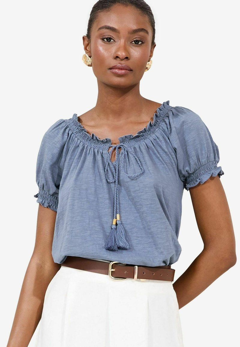 Lichtblauw, kort mouwblouse met gerimpelde halslijn en een trekkoord met kwastjes. Bruine riem gedragen over een witte rok.