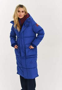 Kobieta w długim, niebieskim pikowanym płaszczu z dużymi kieszeniami i kapturem, nałożonym na bordową kurtkę, stoi na jednolitym tle.