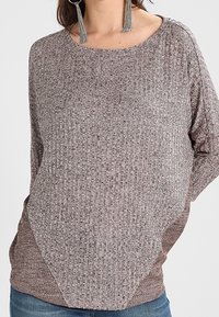 Brun, strukturerad tröja med rundad halsringning och lös passform. Har en geometrisk design vid fållen, ihop med avslappnade blå jeans.