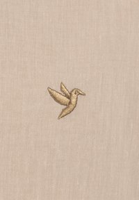 Un tejido beige presenta un pequeño colibrí bordado en oro, ubicado en el centro, con alas extendidas y un pico afilado.