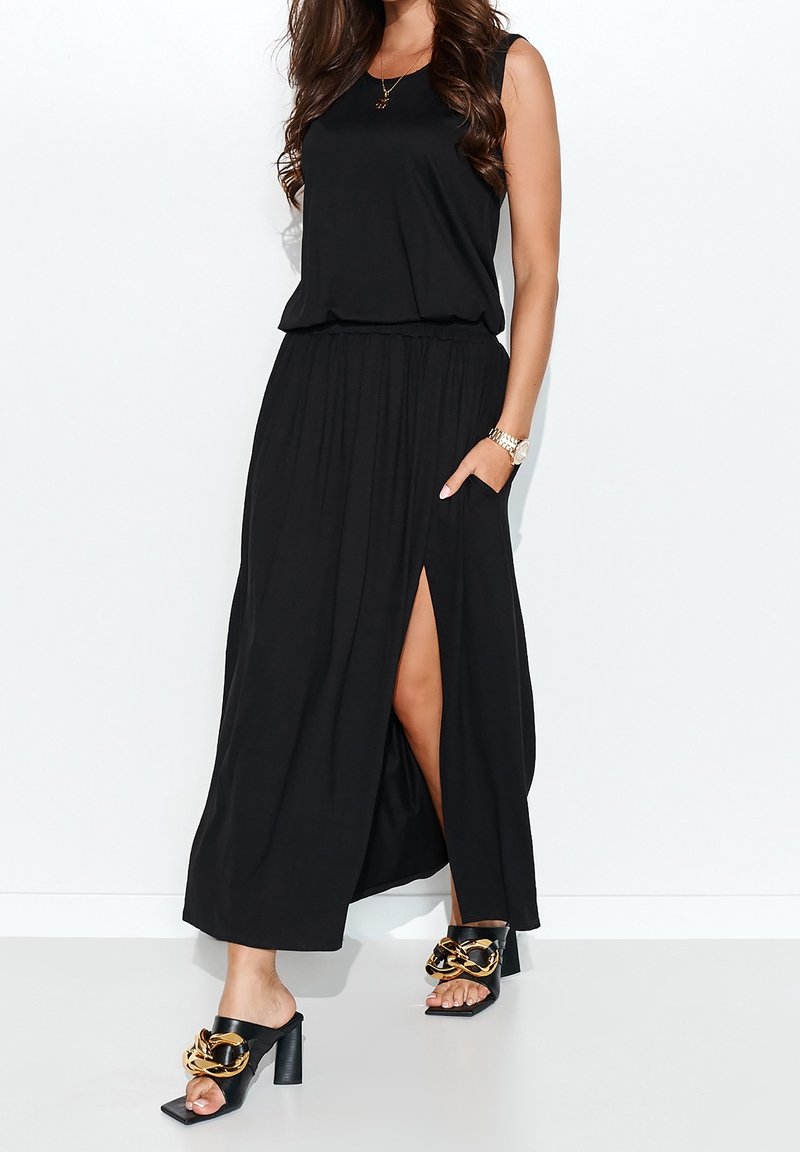 Robe maxi sans manches noire avec une taille froncée, une fente latérale et des poches. Associée à des sandales à talons noires avec des accents dorés.