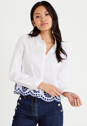 Junge Frau, die den Ärmel eines weißen Hemdes mit blauem besticktem Saum hochkrempelt, trägt dunkelblaue, hoch taillierte Jeans mit seitlichen Knöpfen.