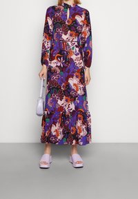 Lila kukallinen mekko, jossa on pitkät hihat, avaimenreikäyksitys kaulassa, vyötäröstä kireä ja röyhelöhelma. Yhdistetty vaalean lilan värisiin korkokenkiin.