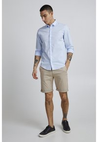 Camisa de botones azul claro, pantalones cortos beige enrollados, zapatillas negras, tatuajes en los brazos, de pie contra un fondo neutro.