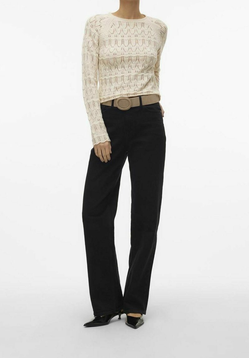 Personne portant un haut à manches longues en maille crème, une ceinture beige avec une boucle ronde, un pantalon large noir et des chaussures pointues noires.