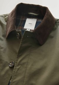 Veste vert olive avec col en velours côtelé marron, fermeture à boutons et doublure en tissu à carreaux visible à l'intérieur du col.