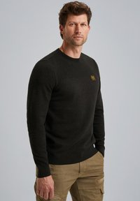 Donker getextureerde trui met een ronde halslijn en een klein goudkleurig logo op de borst,Combineert met beige cargobroek, tegen een neutrale achtergrond.