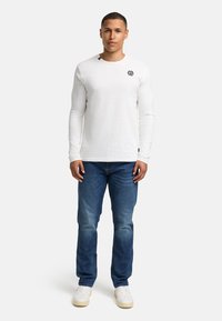 Langermet hvit T-skjorte med svart logo på brystet, kombinert med blå denimjeans og hvite joggesko. Enkelt, tilpasset design.