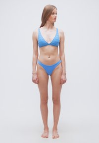 Νεαρή γυναίκα που στέκεται ξυπόλητη φορώντας μπλε υφασμάτινο μπικίνι, κοιτάζοντας προς τα αριστερά της σε απλό λευκό φόντο.