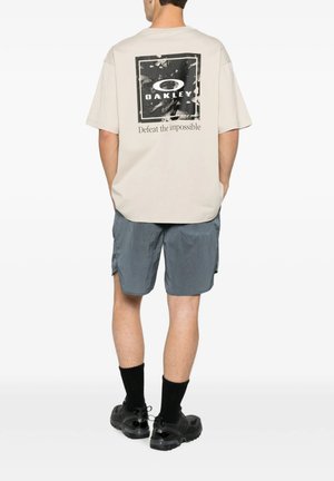 Uomo che indossa una T-shirt beige oversize Oakley con logo e testo, pantaloncini grigi, calzini neri e scarpe da ginnastica nere, in piedi di spalle.