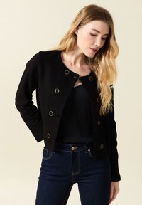 Zwarte cropped jacket met een ronde hals, gouden knopen en lange mouwen; gecombineerd met donkerblauwe jeans. Gladde stoftextuur.