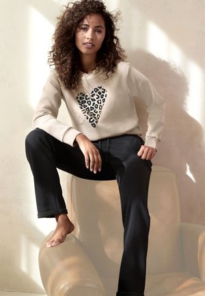 Beige Kapuzenpullover mit einem schwarzen und leopardfarbigen Herzmuster, kombiniert mit schwarzen Hosen. Das Model sitzt auf einem hellen Stuhl vor einem minimalistischen Hintergrund.