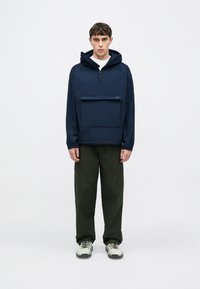 Tengerészkék kapucnis pulóver cipzáros nyakkal és elülső zsebbel, olívazöld zsebes nadrággal és szürke sportcipőkkel fekete talppal párosítva.