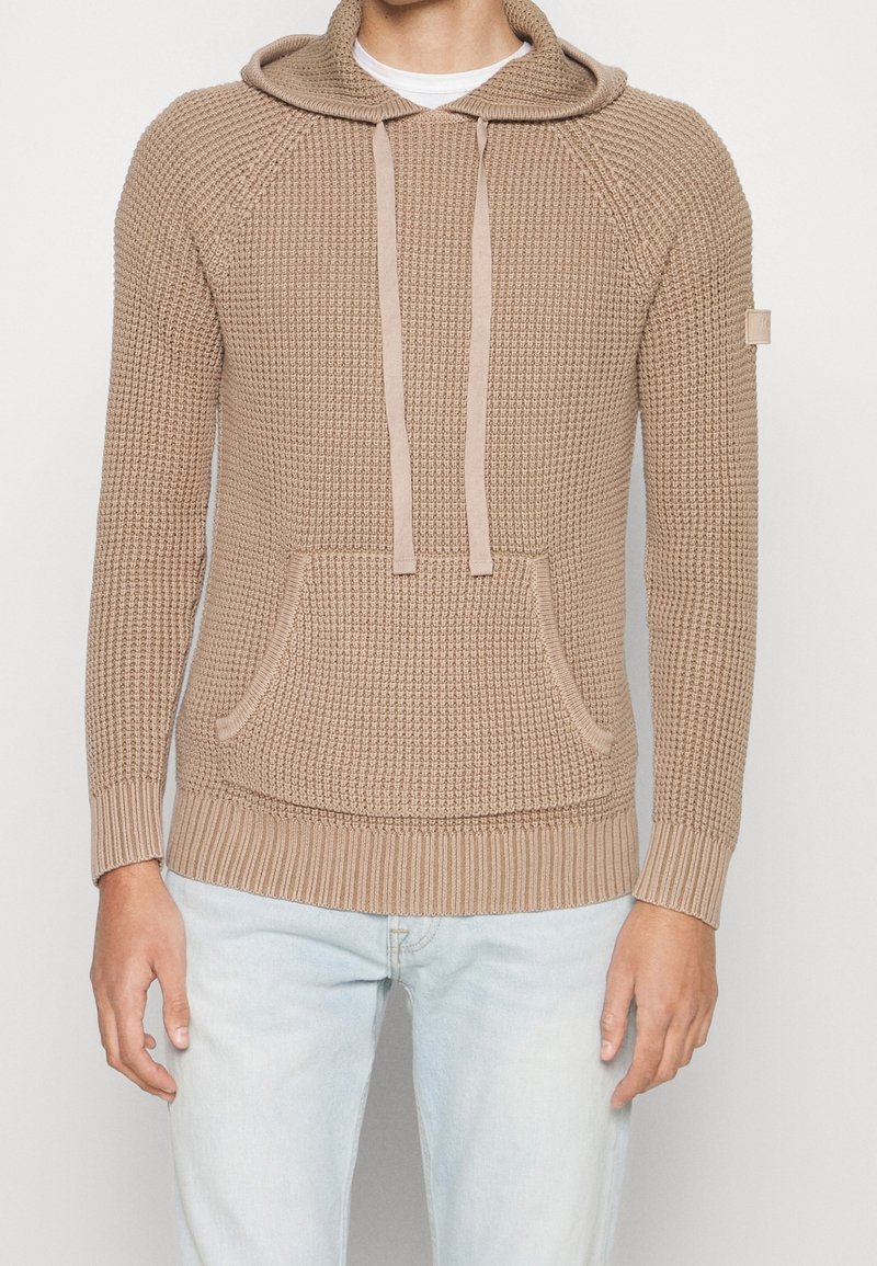 Sweat à capuche beige en maille texturée avec une poche kangourou devant et des cordons, porté sur une chemise blanche et un jean bleu clair.