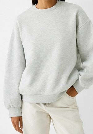 Person wearing a light gray crewneck sweatshirt with long sleeves and off-white pants, one hand in pocket, against a plain background.