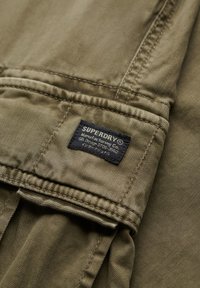 Olivgrüner Stoff mit einer aufgenähten Tasche und einem schwarzen "SUPERDRY"-Etikett mit weißem Text und japanischen Schriftzeichen.