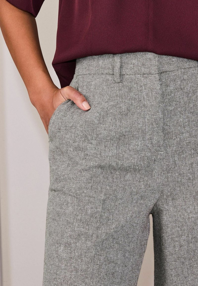 Női kéz a magasderekú, szürke halszálkamintás nadrág elülső zsebében, sötétvörös, betűrt blúzzal párosítva.