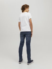 Camiseta blanca de manga corta combinada con jeans azul oscuro. Zapatillas en azul marino con acentos blancos. El modelo se ve desde atrás, girando ligeramente.