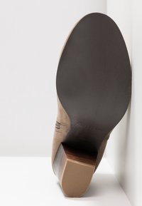 Beżowy zamszowy botek na kostkę z grubym, drewnianym obcasem. Podeszwa jest ciemnobrązowa z teksturowanym wykończeniem. Dodatki obejmują poziome przeszycia.