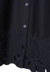 Tissu noir avec boutons à l'avant et broderie florale découpée complexe le long du bord inférieur du vêtement.