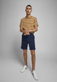 Chemise à manches courtes rayée marron et blanche, shorts bleu marine à revers, baskets blanches assorties avec des accents bleu marine, se tenant contre un fond gris.
