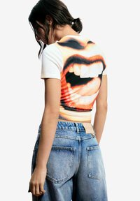 Kurzärmliges, kurz geschnittenes T-Shirt mit lebendigem Grafikdruck eines geöffneten Mundes, weiß an der Vorderseite und farbenfrohem Design auf der Rückseite. Kombiniert mit hoch taillierten blauen Jeans.