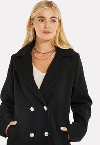 Cappotto nero doppiopetto in tessuto testurizzato, con colletto a revers e bottoni crema. Il modello indossa un top chiaro sotto.