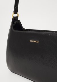 Borsa in pelle nera con superficie texturizzata. Dotata di un logo dorato e di una fettuccia sottile e regolabile fissata con hardware dorato.